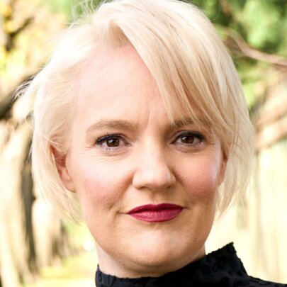 Blonde woman with red lipstick, standing outside under blurred trees.
