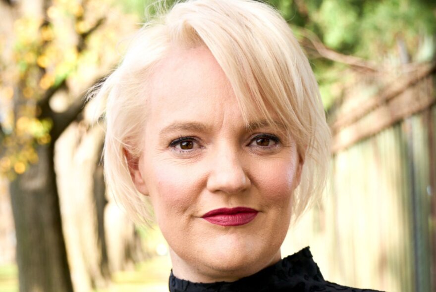 Blonde woman with red lipstick, standing outside under blurred trees.