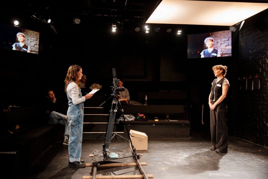 An 11-year old girl in overalls on stage with a woman in dark pants and vest, with a camera on a tripod.