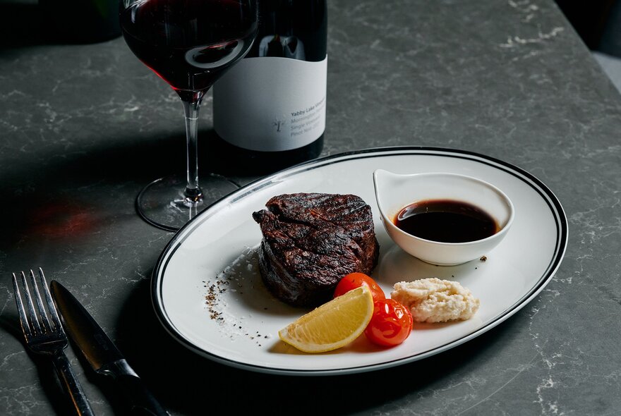 A bone-in steak on a white plate with sauce, cherry tomatoes, and a lemon wedge, next to a glass of red wine.