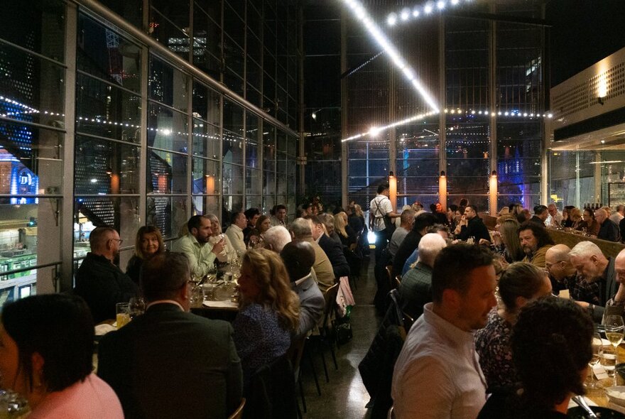 Dining patrons eating and drinking, seated at long tables inside Taxi Kitchen restaurant, with large windows surrounding them and views of the city.