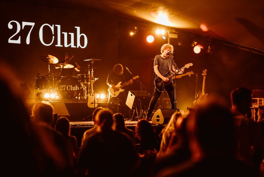 A band playing live on stage, in front of an audience, with the words 27 Club written on the backdrop behind the stage.