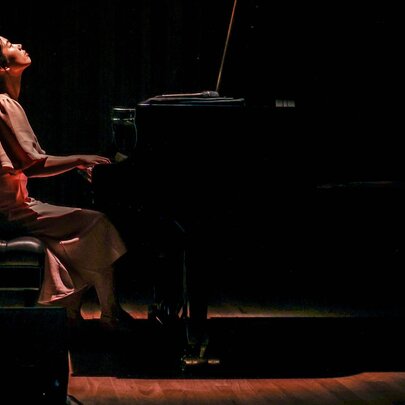 Pianist Mina Yu playing a grand piano while looking up at the ceiling, a single warm light cast down on her. 