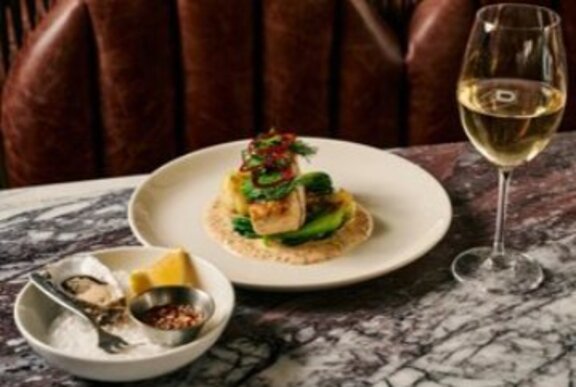 Dish of oysters and plate of food with glass of wine on a marble table.