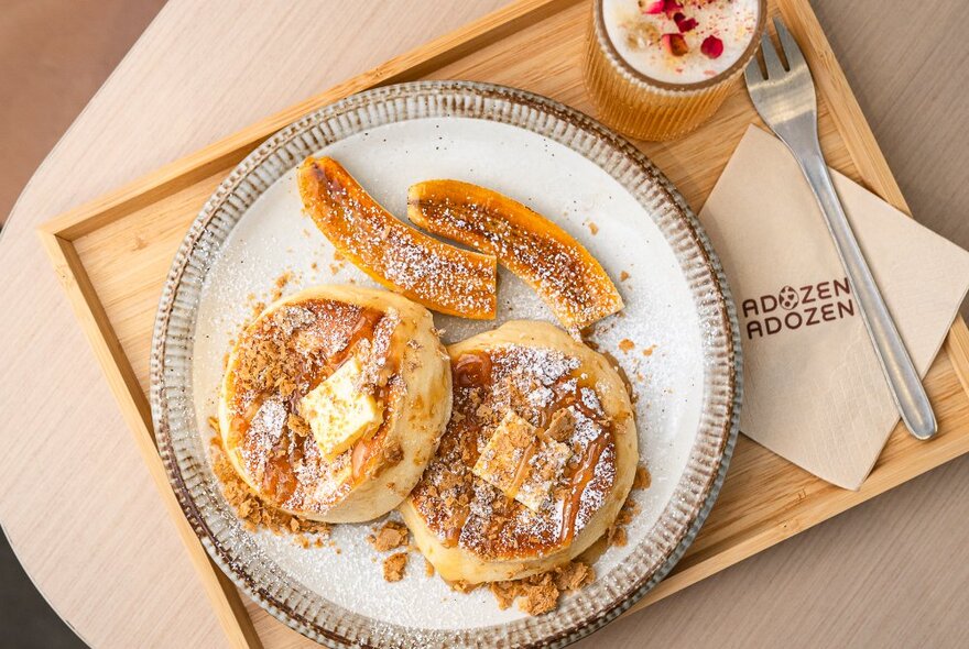 Souffle-style fluffy pancakes with caramelised banana and caramel sauce on a round plate alongside a fork, napkin and drink on a wooden tray.