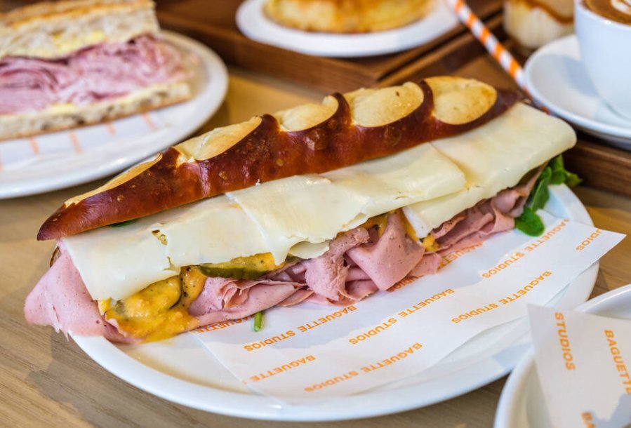A dark brown baguette sandwich filled with deli meat and cheese on a white plate
