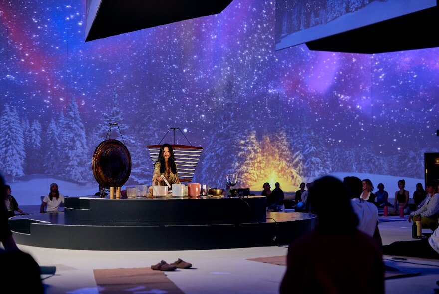 A wellness exercise class seated on the floor in front of a woman seated at a central dais in a wintery backdrop of purple snowy trees.