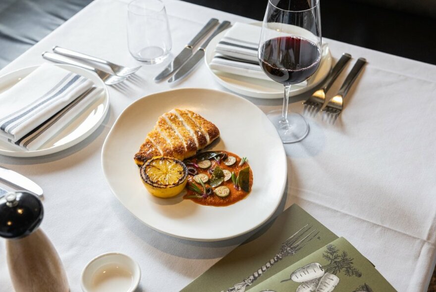 A formal table setting with a plate of food with charred lemon garnish and red wine.