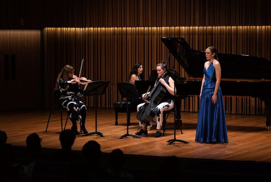 Three musicians playing violin, cello and piano, with singer wearing long blue gown, performing on dimly lit curtained stage.