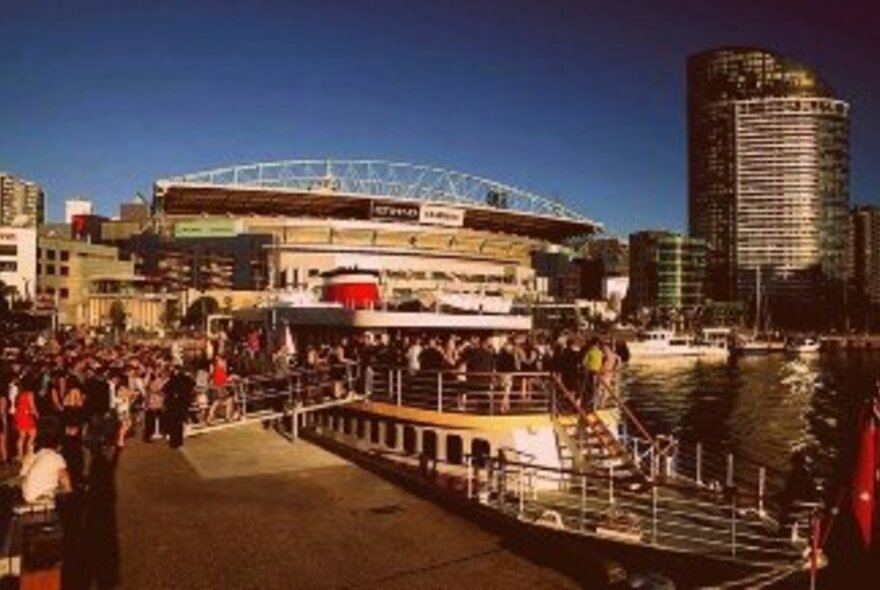 A boat docked at Victoria Harbour Docklands, with people lining up to board, a large sports stadium and a few city buildings visible in the background.