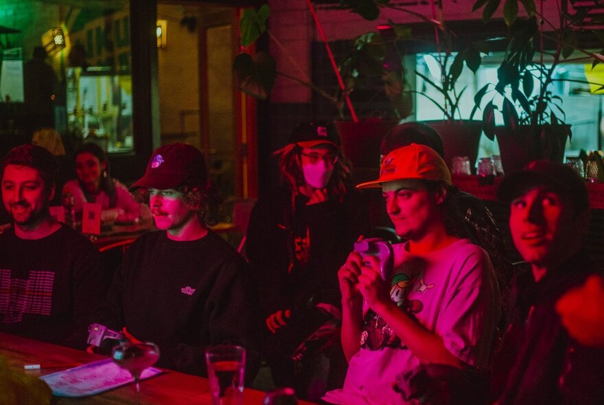 Video game lovers playing a video game in a darkened bar, bathed in pink neon light.