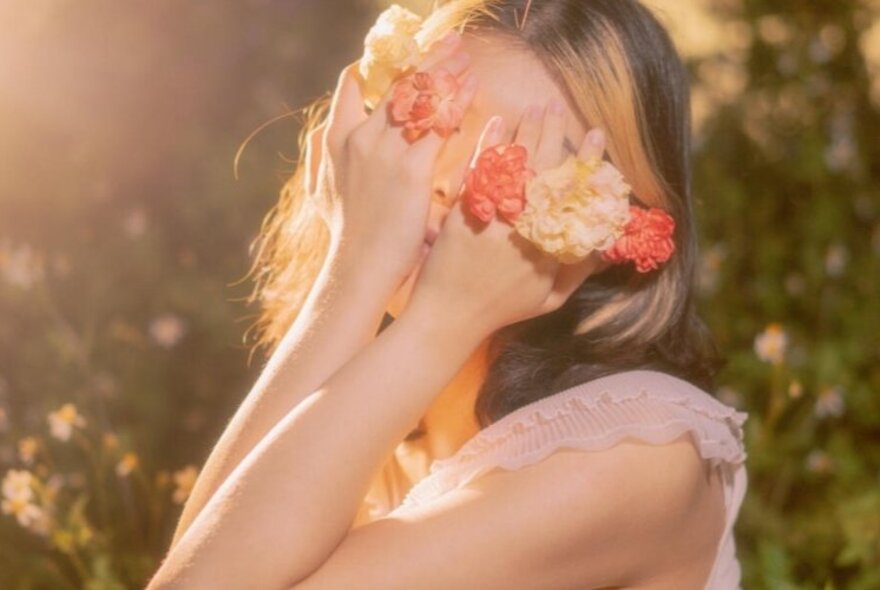 A sunlit portrait of a young woman covering her eyes with her flower-covered hands.