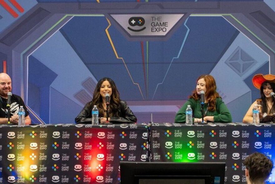 A panel of four people at a gaming expo with coloured lights beneath them. 
