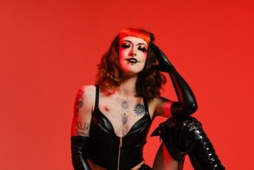 A burlesque performer wearing a black leather vest and black gloves, with tattoos and piercings, posing against a red background.