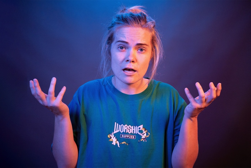A performer with their hands raised in front of their chest, wearing a loose blue t-shirt, with a slightly confused expression on their face.