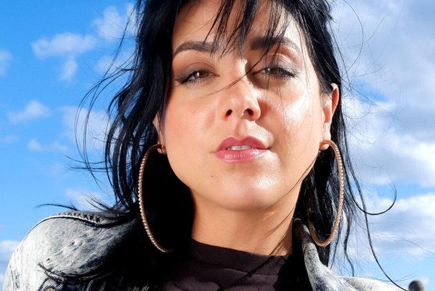 Comedian Bianka Ismailovski with shoulder length dark hair, large hoop earrings and gazing straight at the viewer, the sky with white clouds behind her head. 