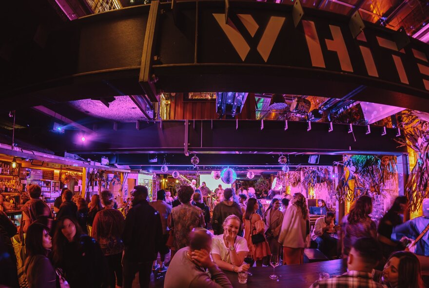 A crowded bar with a pink light cast over the patrons and a large sign that says WHIT... overhead. 