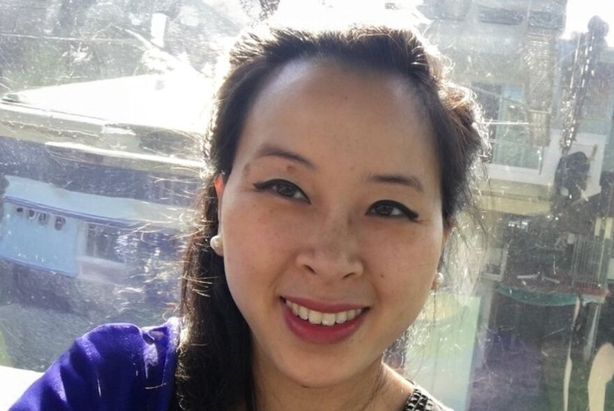 Woman with an open smile standing in front of a window. her long black hair pulled away from her face in a pony tail.