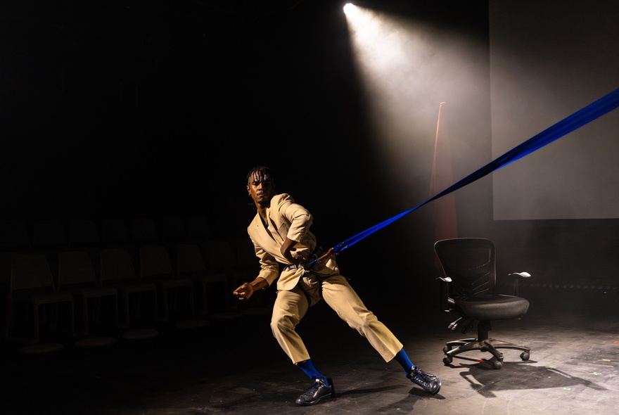 Dancer wearing a beige suit, holding a blue strap in an indoor space.