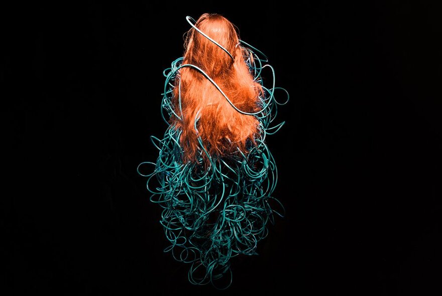 A woman with long red hair viewed from behind, her body encased in blue ropes.