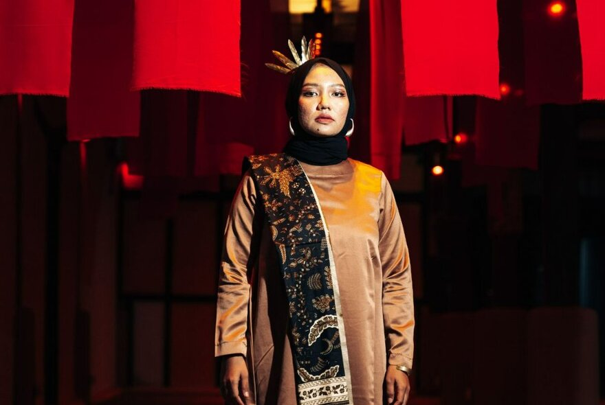 Woman standing and looking directly ahead, wearing a long-sleeved tan coloured dress with an embroidered sash over her shoulder and a headscarf.  