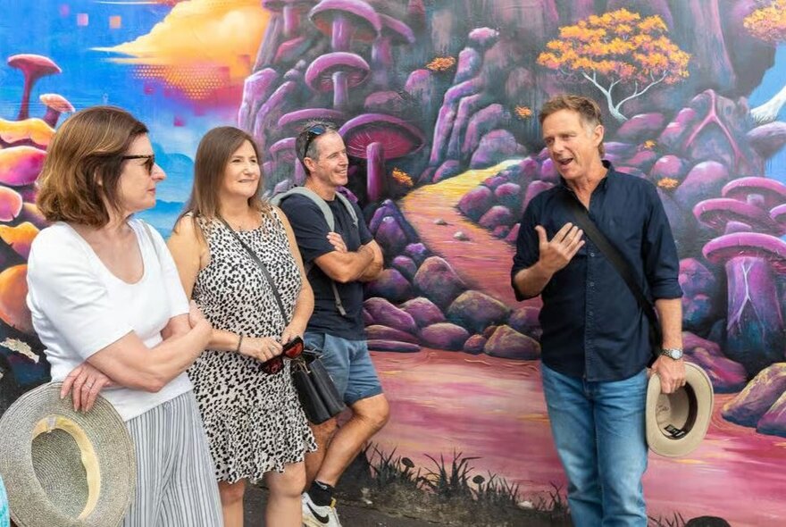 Small tour group listening to a man talking, holding a hat and standing in front of a wall mural.