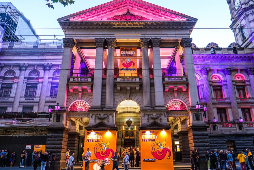 Outside the Melbourne Town Hall with Comedy Festival banners hung out the front. It is lit up in purple and pink lights.