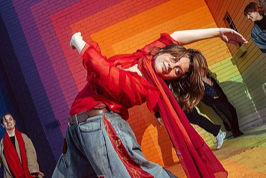 A woman bending backwards in a dance move, her red scarf flowing behind her, in front of a rainbow-coloured brick wall.