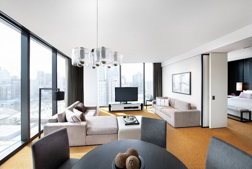 Hotel room with orange carpet and grey furnishings, separate lounge area with seating and TV opening to doorway to bedroom.