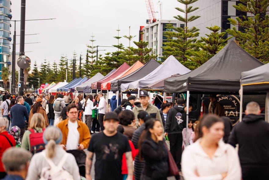 People walking past rows of outdoor market stalls with umbrellas and trees.