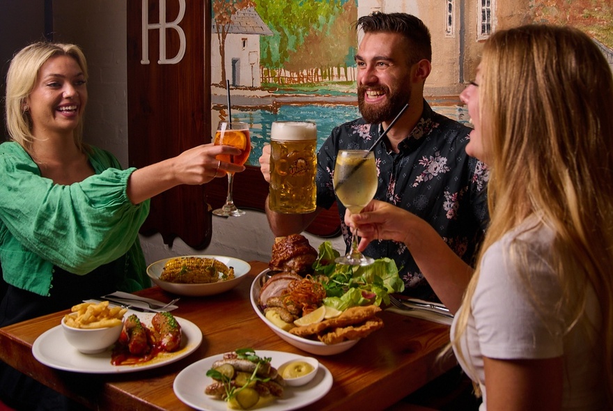 Three friends seated around a table raising their glasses with plates of food in front of them.