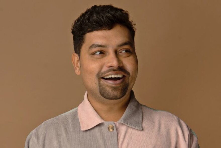 Portrait of comedian, Navin Noronha, in front of a brown background.