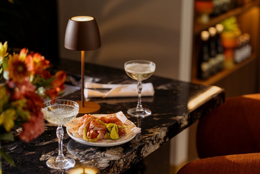 Two cocktail glasses and a plated Italian appetizer on a dark marble restaurant bar counter under warm lighting.