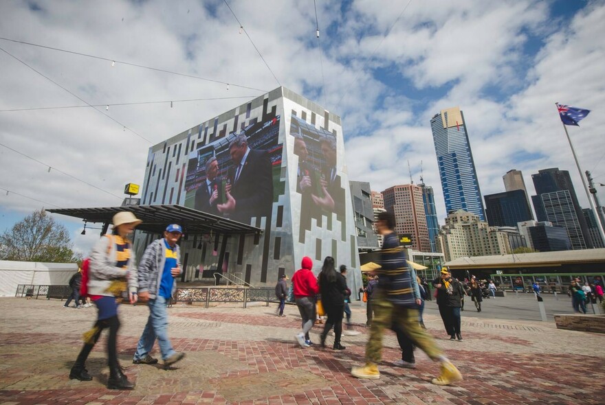 People walking through Fed Square, past large outdoor screen.