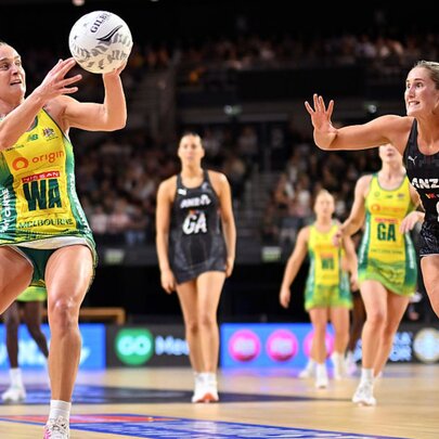 A netball player in a green and gold uniform catches the ball while a defender in a black uniform lunges toward her on an indoor court, blurred players and seated fans in the background.