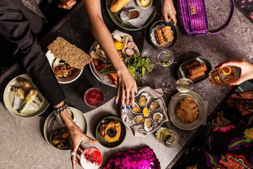 A table heaving with share plate dishes and drinks and three arms reaching out to pick up some of the offerings. 