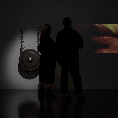 Installation view of two artworks hanging in a dimly lit space in a gallery, with the silhouette of two people standing in front of the works.
