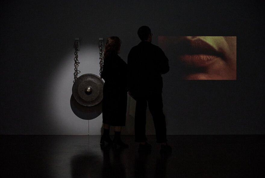 Installation view of two artworks hanging in a dimly lit space in a gallery, with the silhouette of two people standing in front of the works.