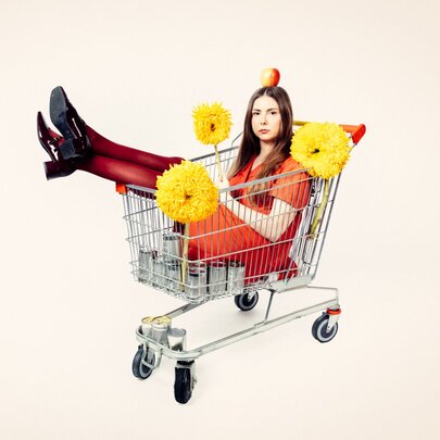 A woman wearing an orange dress, an apple  balanced on her head, sitting inside a metal shopping trolley with her legs pointing up over the edge and three large, yellow pom-poms stuck to the trolley.