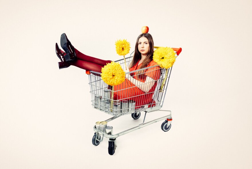 A woman wearing an orange dress, an apple  balanced on her head, sitting inside a metal shopping trolley with her legs pointing up over the edge and three large, yellow pom-poms stuck to the trolley.