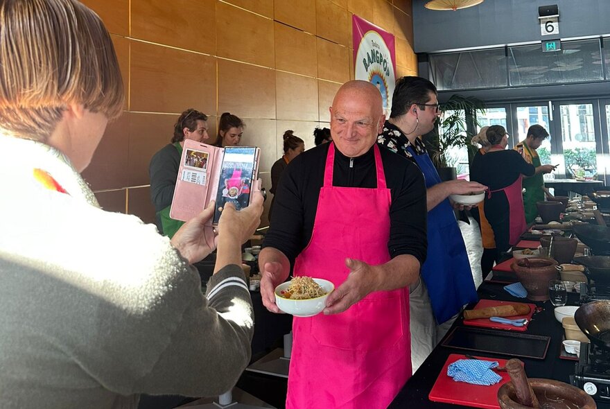 Man wearing a bright pink outfit holding a bowl of food, being photographed on a mobile phone.