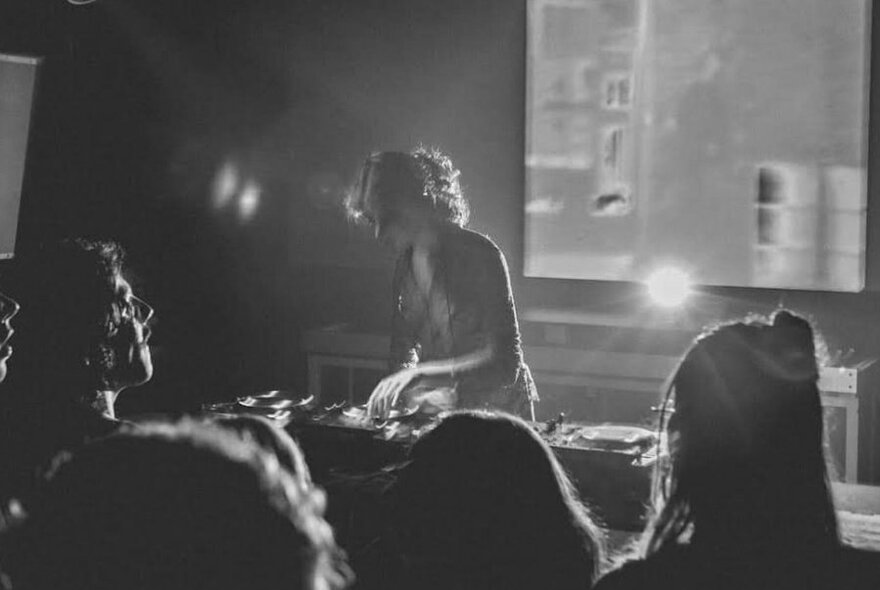 A DJ playing on a mixing desk, with a screen behind them and close-ups of the back of people's heads watching the DJ on stage.