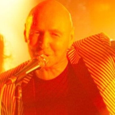 Man with no hair singing into a microphone live on stage, another person on stage in the background, the entire image bathed in a warm yellow-orange light.