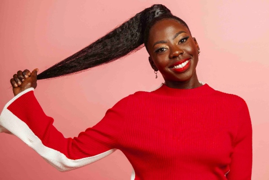 Smiling comedian pulling her ponytail out with one hand, wearing a red and white bell-sleeved top.