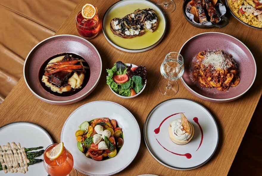 Looking down at a selection of dishes and bowls of food, and beverages in glasses, on a wooden table top.