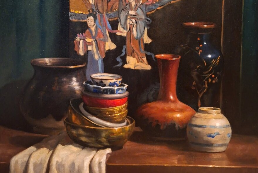 Still life oil painting of a stack of ceramic bowls assorted pottery vases, fabric and a painting, all resting on a wooden sideboard. 
