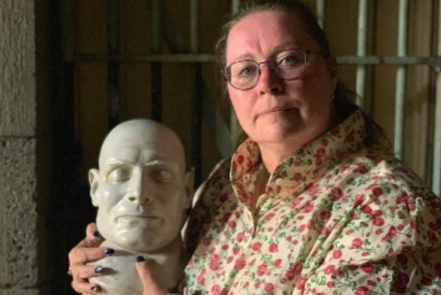 Woman with spectacles holding the death mask of Ned Kelly in an old jail cell.