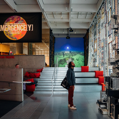 A person is inside a modern art gallery looking up at a towering sculpture behind them is a large banner saying emergency 