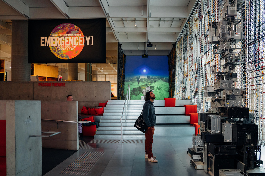 A person is inside a modern art gallery looking up at a towering sculpture behind them is a large banner saying emergency 
