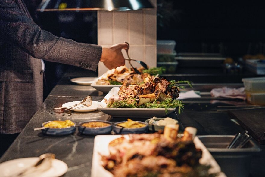 A view looking down a long table at a buffet wtih a hand selecting food from a plate with tongs. 